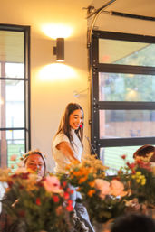 Smiling presenter with a headset in a bright modern event space with glass-panel doors, interacting with seated guests at a table filled with colorful floral arrangements.