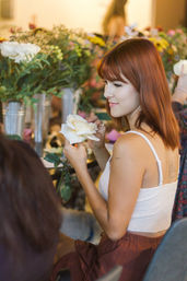 Red-haired woman holding a white rose and arranging flowers at an indoor floral workshop with metal vases and mixed bouquets