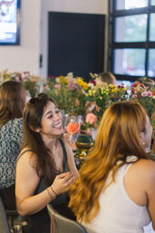 Women laughing and chatting at an indoor, flower-filled table with colorful bouquets and vases in a cozy event space.