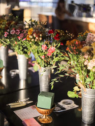 Indoor flower-arranging station with colorful mixed bouquets in metal vases, green floral foam block on a small gold pedestal, scissors and tape on a black table with soft cafe-style background