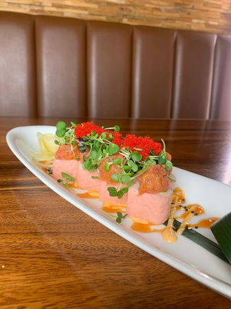Close-up of vibrant sushi rolls on a white rectangular plate: pink soy-wrapped pieces topped with spicy tuna, bright red tobiko and microgreens, drizzled with spicy mayo and eel sauce on a wood table in a casual sushi restaurant booth.