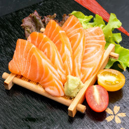 Fresh salmon sashimi slices fanned on a small bamboo mat with wasabi, lettuce and halved cherry tomatoes on a black serving tray, Japanese cuisine
