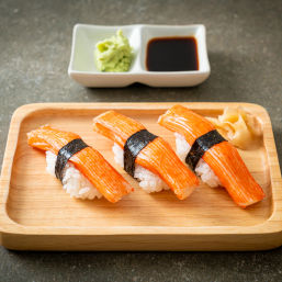 Three bright orange kani (imitation crab) nigiri sushi on a wooden tray, each wrapped with nori and served with pickled ginger and a dish of soy sauce and wasabi.