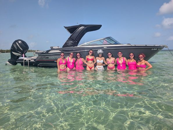 Group of women in pink swimsuits and sunglasses posing waist‑deep in clear turquoise shallow water beside a sleek black motorboat under a sunny blue sky.