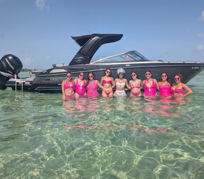 Group of women in pink swimsuits and sunglasses posing waist‑deep in clear turquoise shallow water beside a sleek black motorboat under a sunny blue sky.