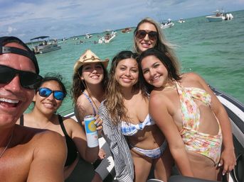 Group selfie of six people in swimwear on a motorboat in clear turquoise coastal waters, smiling under a sunny sky with anchored boats nearby — relaxed beach day vibe.