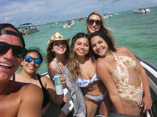 Group selfie of six people in swimwear on a motorboat in clear turquoise coastal waters, smiling under a sunny sky with anchored boats nearby — relaxed beach day vibe.