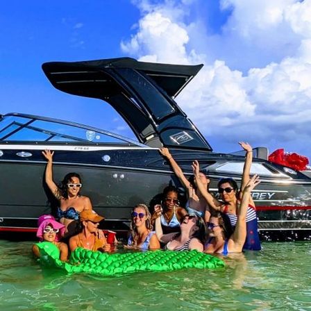 Group of friends enjoying a lively boat party in shallow turquoise water beside a sleek black speedboat, lounging on a bright green inflatable under a sunny blue sky.