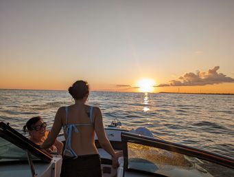 Two people on a motorboat at sunset — one standing at the stern in a blue bikini top, the other seated with sunglasses — golden sun reflecting across calm ocean and orange sky.