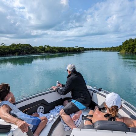 Three people lounging on a motorboat cruising a calm turquoise, mangrove-lined inlet under a bright, partly cloudy sky.