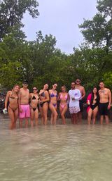 Group of friends in colorful swimsuits standing in shallow clear water along a tropical mangrove shoreline, posing for a smiling beach hangout under an overcast sky.