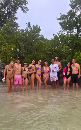Group of friends in colorful swimsuits standing in shallow clear water along a tropical mangrove shoreline, posing for a smiling beach hangout under an overcast sky.