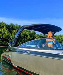 Smiling woman in a blue bikini at the helm of a sleek motorboat anchored in a coastal mangrove inlet under a clear blue sky, holding a travel tumbler.