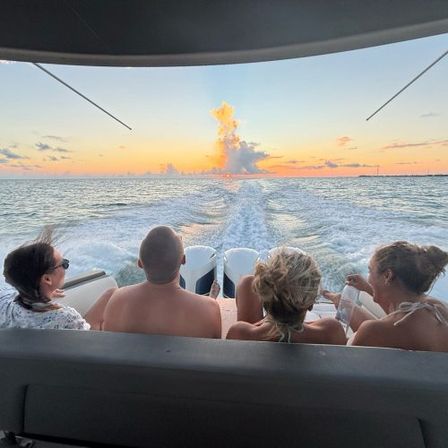 Four people seated on a boat’s stern watching a colorful sunset over calm coastal waters, twin-engine wake trailing behind and a glowing cloud plume on the horizon.