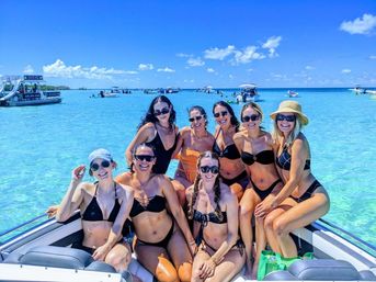 Cheerful group of eight women in swimsuits posing on a boat bow over shallow turquoise tropical water, with other boats and a bright blue sky in the background.