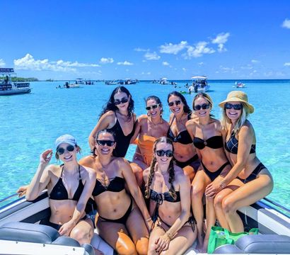 Cheerful group of eight women in swimsuits posing on a boat bow over shallow turquoise tropical water, with other boats and a bright blue sky in the background.