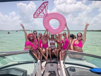 Friends in pink swimsuits posing on a boat in turquoise tropical water under a sunny sky, holding a giant pink inflatable ring float and celebrating.