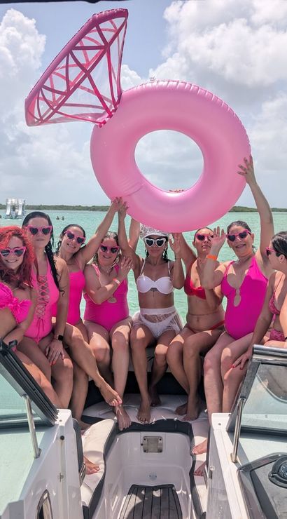 Friends in pink swimsuits posing on a boat in turquoise tropical water under a sunny sky, holding a giant pink inflatable ring float and celebrating.