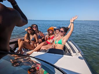 Group of five friends in colorful bikinis with sunglasses and sun hats, laughing and holding drinks at the bow of a motorboat on calm coastal bay waters under a clear summer sky.