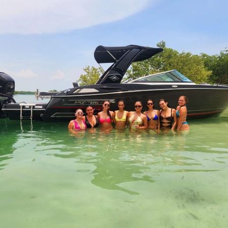 Eight people in colorful bikinis wading in clear shallow green water beside a sleek black motorboat under a sunny sky — summer boat party