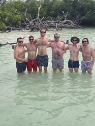 Six friends standing waist-deep in shallow turquoise water by a tropical mangrove shoreline, shirtless in swim trunks and sunglasses, posing with drinks on a sunny beach day.