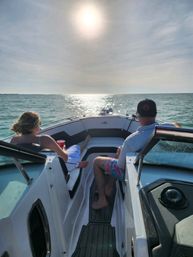 Two people relaxing on the bow of a motorboat as the sun sparkles on open water toward the horizon, one holding a red cup and the other wearing a cap and colorful shorts.