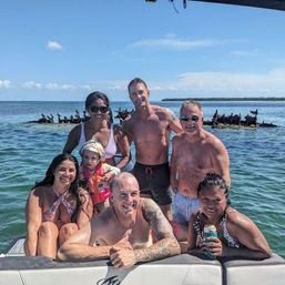 Group of friends and family enjoying a boat day in tropical turquoise coastal waters, with old wooden pilings and seabirds under a sunny blue sky.