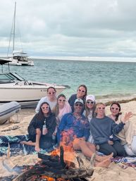 Eight friends wearing heart-shaped sunglasses gathered on a sandy beach around a bonfire with turquoise sea and an anchored sailboat under a cloudy sky