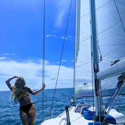 Person with long blonde hair in a black bikini standing on a sailboat deck, holding a rope and gazing across the blue ocean under a bright sunny sky with full white sails.