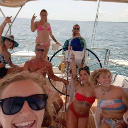 Smiling group of friends in bikinis partying on a sailboat in sunny open water, steering wheel and drinks visible