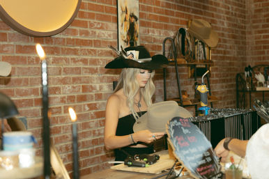Blonde artisan examining a cowboy hat at a rustic brick-walled western hat shop counter