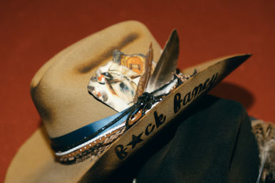 Tan felt cowboy hat with braided leather band, patterned scarf and feathers tucked in; black cursive writing on the brim, photographed on a warm red background.