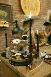 Rustic boutique counter with a tiered wooden tray of ribbon spools, amber candlesticks holding lit black taper candles, a black desk lamp and cowboy hats displayed against an exposed brick wall — crafty western boutique vibe.