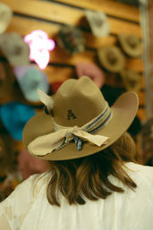 Rear view of a person wearing a tan felt cowboy hat stamped with an "A" and tied with a frayed fabric band, standing in front of a wall of hats in a rustic shop