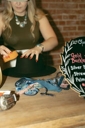 Hands with red nails cleaning a black wide-brim hat with a sponge on a wooden table, fabric ribbons and a chalkboard sign nearby in a cozy boutique display.
