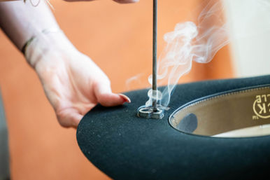 Hand holding a black felt hat on a hat block while a steam wand shapes the brim, white steam curling upward — millinery hat-making scene