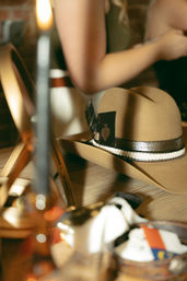 Brown felt western hat with decorative ribbon and an ace of spades tucked in the band, resting on a wooden vanity with a blurred mirror and person adjusting clothing in the background.