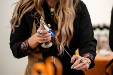 Close-up of artisan using a handheld blowtorch with a blue flame to heat a metal rod for jewelry-making in a craft studio, hands with turquoise rings and painted nails