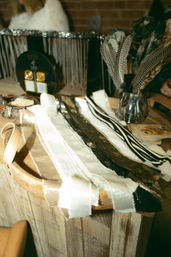 Boho craft display on a rustic wooden tray: neutral fabric swatches, metallic trims, striped ribbons and decorative feathers in a small glass jar