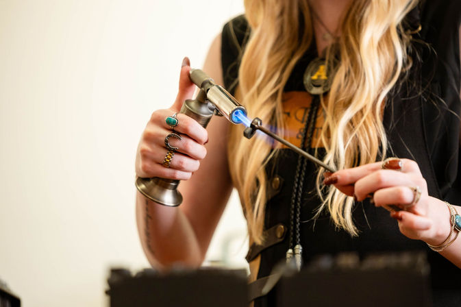 Close-up of artisan hands using a blue-flame torch to heat a metal rod for jewelry making, hands adorned with rings and long blonde hair in a studio workshop