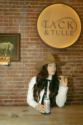 Woman in a cowboy hat at a rustic brick-walled bar, holding a cigar and a can of Coors Light beneath a round illuminated wall sign.