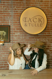 Two friends trying on brown cowboy hats at a wooden counter against a red brick wall, smiling beneath a round backlit wall sign in a western-style shop