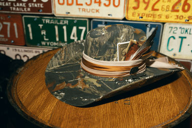 Camouflage cowboy hat with a ribbon band, feathers and tucked playing cards, resting on a wooden barrel table in front of a wall of vintage license plates — rustic Western decor.
