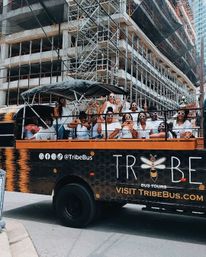 Group of people in white outfits and matching round sunglasses cheering on an open-top tour bus rolling past a downtown high-rise under construction