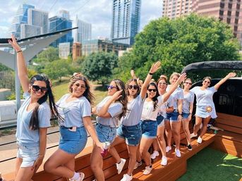 Group of women in white tees and denim shorts posing and waving on a rooftop deck with an urban skyline and park trees on a sunny day