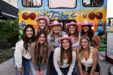 Eight smiling friends in pink cowboy hats pose cheerfully in front of a colorful bus painted with "Tribe" on a city street, playful group photo vibe.