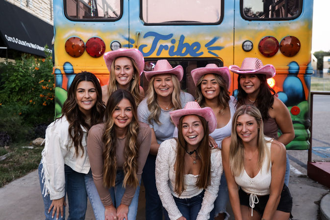 Eight smiling friends in pink cowboy hats pose cheerfully in front of a colorful bus painted with "Tribe" on a city street, playful group photo vibe.