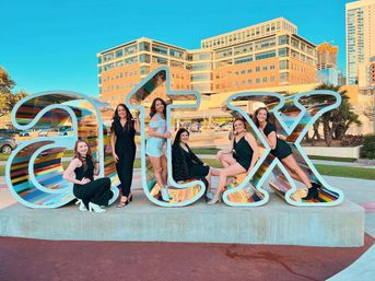 Six women posing on and around a large rainbow-striped "ATX" public art sign in an urban plaza with modern buildings and a bright blue sky