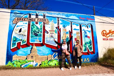 Two people posing in front of a colorful "Greetings from Austin" mural featuring the Texas State Capitol, skyline scenes and a Texas flag on a sunny day