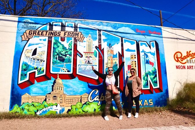 Two people posing in front of a colorful "Greetings from Austin" mural featuring the Texas State Capitol, skyline scenes and a Texas flag on a sunny day